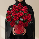 A bouquet of deep red roses with green leaves arranged in a red cylindrical container with black accents, displayed on a round white pedestal against a warm beige background with soft window shadows.