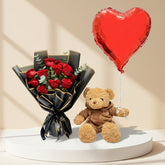 A person in a black robe holds a bouquet of red roses wrapped in black and gold paper, alongside a small brown teddy bear wearing a brown jacket, with a large red heart-shaped foil balloon obscuring the face. The background is a softly lit beige interior with arched alcoves and floral arrangements.