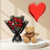 A person in a black robe holds a bouquet of red roses wrapped in black and gold paper, alongside a small brown teddy bear wearing a brown jacket, with a large red heart-shaped foil balloon obscuring the face. The background is a softly lit beige interior with arched alcoves and floral arrangements.