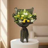 A person in a black outfit and headscarf holds a large bouquet of yellow and white flowers wrapped in dark gray paper with a small card inside a softly lit indoor setting.