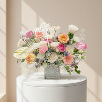 A vibrant floral arrangement in a square glass vase filled with white pebbles, featuring pink and white roses, orange gerbera daisies, pink anthuriums, white carnations, and green foliage, with a white script 'Happy' sign in the center, set against a solid white background.