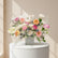 A vibrant floral arrangement in a square glass vase filled with white pebbles, featuring pink and white roses, orange gerbera daisies, pink anthuriums, white carnations, and green foliage, with a white script 'Happy' sign in the center, set against a solid white background.
