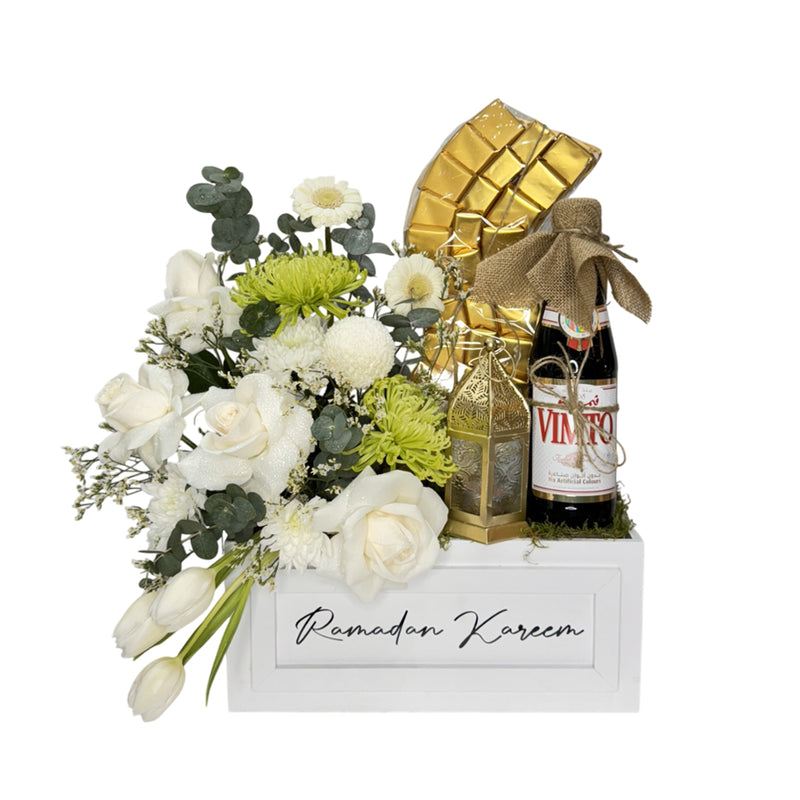 An elegant Eid gift box with fresh white and green flowers, a golden chocolate arrangement, a lantern, and a Vimto bottle—perfect for festive gifting.