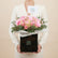 A bouquet of pink roses in a black box with white tissue paper, green foliage, and a small card reading Viva Flora, displayed on a white pedestal against a beige background.