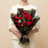 A bouquet of 15 red roses wrapped in black paper with gold edges and a black ribbon, displayed on a white cylindrical pedestal against a beige background with soft shadows.