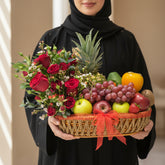 A woven basket gift arrangement filled with red roses, green and red apples, purple grapes, a pineapple, an orange, a kiwi, and strawberries, tied with a red ribbon and displayed on a white pedestal.