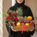 A woven basket gift arrangement filled with red roses, green and red apples, purple grapes, a pineapple, an orange, a kiwi, and strawberries, tied with a red ribbon and displayed on a white pedestal.