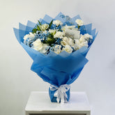 A bouquet of white roses, blue baby's breath, and green foliage wrapped in bright blue tissue paper and tied with a white ribbon, displayed on a white pedestal against a light gray background.