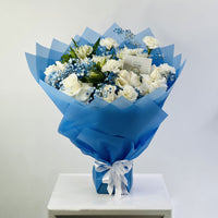 A bouquet of white roses, blue baby's breath, and green foliage wrapped in bright blue tissue paper and tied with a white ribbon, displayed on a white pedestal against a light gray background.