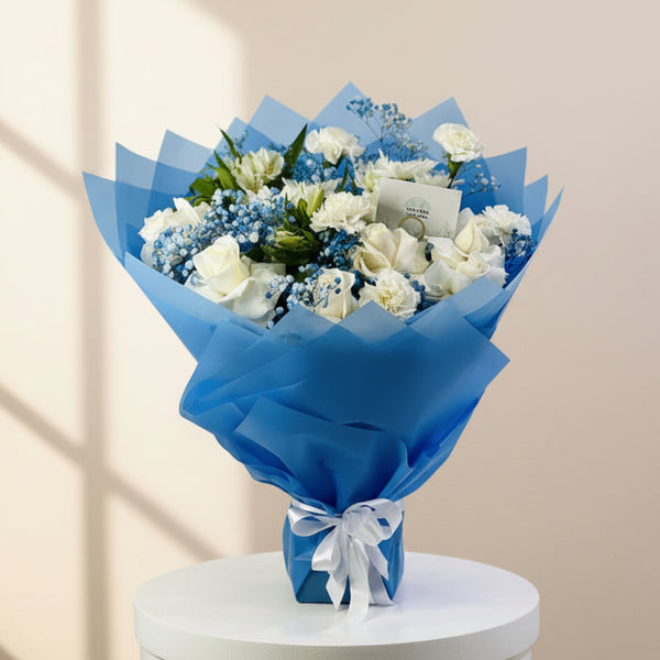 A bouquet of white roses, blue baby's breath, and green foliage wrapped in bright blue tissue paper and tied with a white ribbon, displayed on a white pedestal against a light gray background.