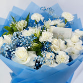 A bouquet of white roses, carnations, and baby's breath flowers wrapped in light blue tissue paper, with a small card tucked inside.