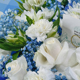 A close-up of a floral arrangement featuring white roses, white carnations, blue baby's breath, and green leaves, with a white card partially visible on the right.