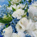 A close-up of a floral arrangement featuring white roses, white carnations, blue baby's breath, and green leaves, with a white card partially visible on the right.