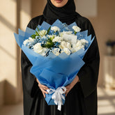 A bouquet of white roses, blue baby's breath, and green foliage wrapped in bright blue tissue paper and tied with a white ribbon, displayed on a white pedestal against a light gray background.