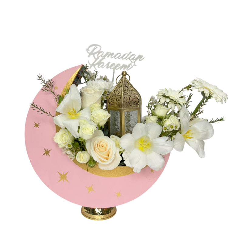 A crescent moon-shaped floral arrangement with white roses, lilies, and a decorative lantern, set against a pink and gold background with 