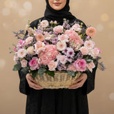 A vibrant artificial flower arrangement in a woven wicker basket, featuring pink, peach, and purple blooms with green foliage, displayed on a white cylindrical pedestal against a warm beige background.