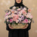 A vibrant artificial flower arrangement in a woven wicker basket, featuring pink, peach, and purple blooms with green foliage, displayed on a white cylindrical pedestal against a warm beige background.