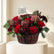 A wicker basket filled with a winter floral arrangement featuring red roses, burgundy protea, green pine branches, and dark burgundy foliage, displayed on a white pedestal against a white wall.