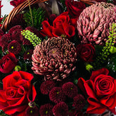 A wicker flower basket filled with red roses, burgundy protea flowers, dark green foliage, and a small white card holder, photographed against a plain light background.