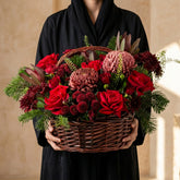 a lady holding a flower basket. A wicker basket filled with a winter floral arrangement featuring red roses, burgundy protea, green pine branches, and dark burgundy foliage,