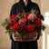 a lady holding a flower basket. A wicker basket filled with a winter floral arrangement featuring red roses, burgundy protea, green pine branches, and dark burgundy foliage,