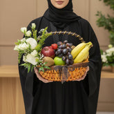 A woven wicker fruit basket filled with bananas, grapes, an apple, a kiwi, and a small red fruit, decorated with white flowers and greenery, displayed on a white pedestal against a beige background.