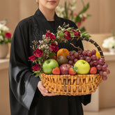 A woven wicker basket filled with fresh fruit and decorated with red roses and green leaves, displayed on a white round pedestal against a warm beige background.