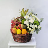 A wicker basket filled with assorted fruits and flowers, placed on a white pedestal against a light gray background. The basket contains red grapes, mangoes, oranges, strawberries, kiwi, an avocado, a pineapple, and white and green flowers.