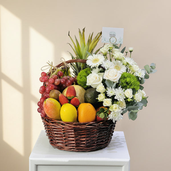 A wicker basket filled with assorted fruits and flowers, placed on a white pedestal against a light gray background. The basket contains red grapes, mangoes, oranges, strawberries, kiwi, an avocado, a pineapple, and white and green flowers.