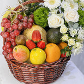 A wicker basket filled with assorted fresh fruits and flowers, including red grapes, strawberries, oranges, a yellow fruit, an apple, kiwi, a melon, green foliage, and white and cream flowers.