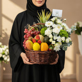 A wicker basket filled with assorted fruits and flowers, placed on a white pedestal against a light gray background. The basket contains red grapes, mangoes, oranges, strawberries, kiwi, an avocado, a pineapple, and white and green flowers.