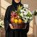 A wicker basket filled with assorted fruits and flowers, placed on a white pedestal against a light gray background. The basket contains red grapes, mangoes, oranges, strawberries, kiwi, an avocado, a pineapple, and white and green flowers.