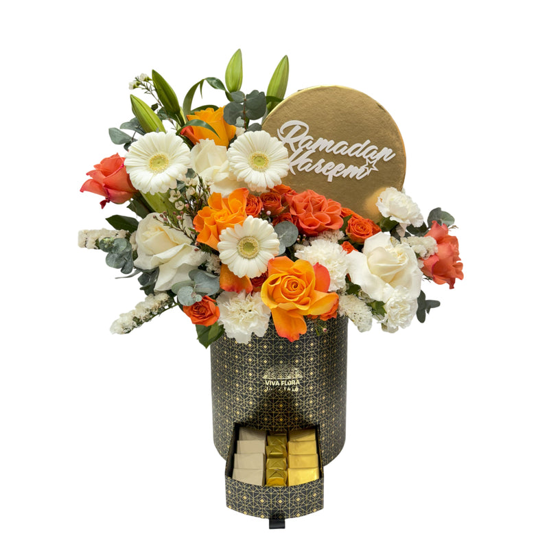 A black and gold floral box with orange and white roses, gerberas, and lilies, featuring a 