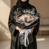 A bouquet of white and light purple roses wrapped in black paper with a light purple inner liner and a white ribbon bow, featuring a silver Arabic nameplate topper.