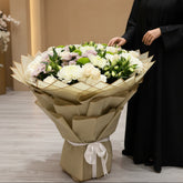 A large floral bouquet wrapped in beige paper and tied with a white ribbon, displayed indoors on a light gray floor with a person standing behind it in a shop setting.