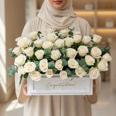 A large bouquet of cream-colored roses with green eucalyptus leaves arranged in a white rectangular box with a gold 'Congratulations!' script, displayed on a white pedestal against a warm beige wall with soft window shadows.