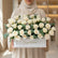 A large bouquet of cream-colored roses with green eucalyptus leaves arranged in a white rectangular box with a gold 'Congratulations!' script, displayed on a white pedestal against a warm beige wall with soft window shadows.