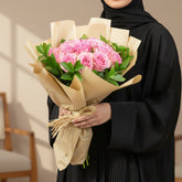 A bouquet of pink roses wrapped in beige kraft paper and tied with a natural twine bow, displayed on a round white pedestal against a warm beige background with soft window-shadow light.