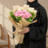 A bouquet of pink roses wrapped in beige kraft paper and tied with a natural twine bow, displayed on a round white pedestal against a warm beige background with soft window-shadow light.