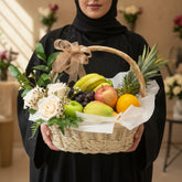 A woven wicker fruit basket filled with fresh fruit and flowers, tied with a beige ribbon and a small branded card, displayed on a white pedestal against a warm beige background.