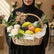 A woven wicker fruit basket filled with fresh fruit and flowers, tied with a beige ribbon and a small branded card, displayed on a white pedestal against a warm beige background.