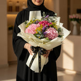 A graduation-themed flower bouquet wrapped in cream paper with a black ribbon printed with 'graduation' in gold, featuring pink, purple, and yellow flowers, displayed on a white pedestal against a beige background.