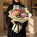 A graduation-themed flower bouquet wrapped in cream paper with a black ribbon printed with 'graduation' in gold, featuring pink, purple, and yellow flowers, displayed on a white pedestal against a beige background.