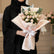 A bouquet of pale pink roses wrapped in blush pink paper with a white ribbon, displayed on a white pedestal against a warm beige background with soft window-shadow lighting.