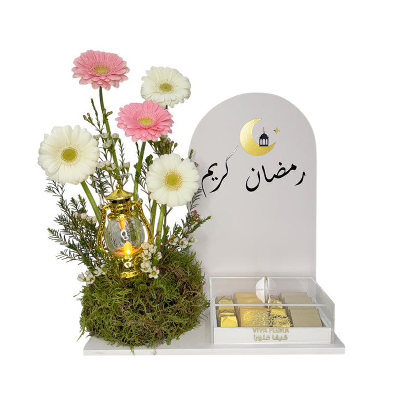 A Ramadan floral arrangement with pink and white gerbera daisies, a gold lantern, and a white sign with 'Ramadan Kareem' in Arabic, next to a box of chocolates.