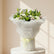 A large floral bouquet wrapped in white tissue paper and tied with a pale yellow ribbon, placed on a light gray floor in front of a glass door. The bouquet contains white and pale pink flowers with green foliage.