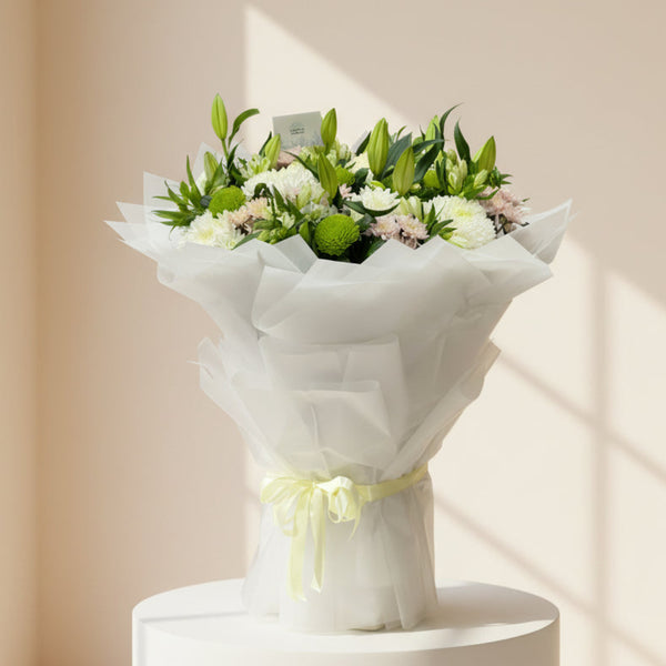 A large floral bouquet wrapped in white tissue paper and tied with a pale yellow ribbon, placed on a light gray floor in front of a glass door. The bouquet contains white and pale pink flowers with green foliage.