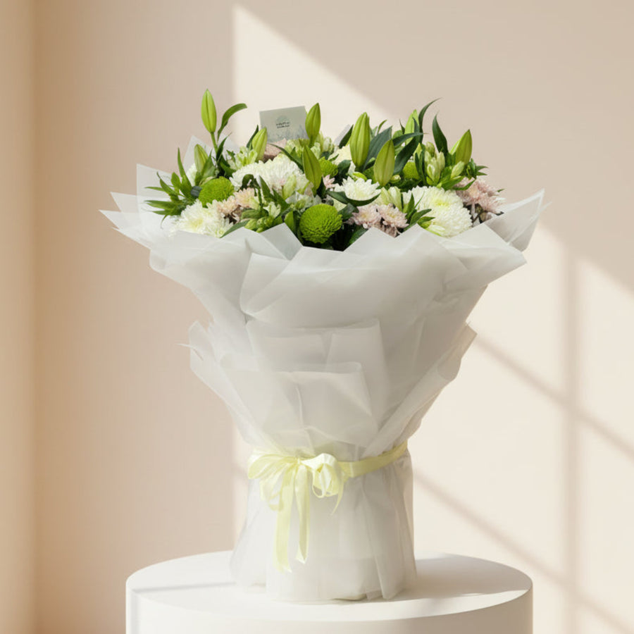 A large floral bouquet wrapped in white tissue paper and tied with a pale yellow ribbon, placed on a light gray floor in front of a glass door. The bouquet contains white and pale pink flowers with green foliage.