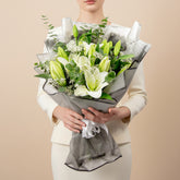A bouquet of white lilies and green buds wrapped in gray paper with a white ribbon, displayed on a white pedestal against a warm beige background.