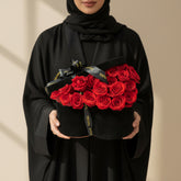 A black cylindrical gift box filled with bright red roses and tied with a black ribbon printed with gold lettering, displayed on a white cylindrical pedestal against a warm beige wall with soft window-shadow lighting.